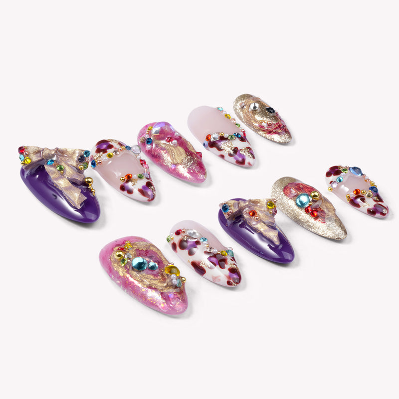 A flat lay of ten almond-shaped press-on nails on a white background featuring deep purple and pearly pink finishes, decorated with colorful rhinestones, pearls, 3D purple bows, and molten gold metallic accents
