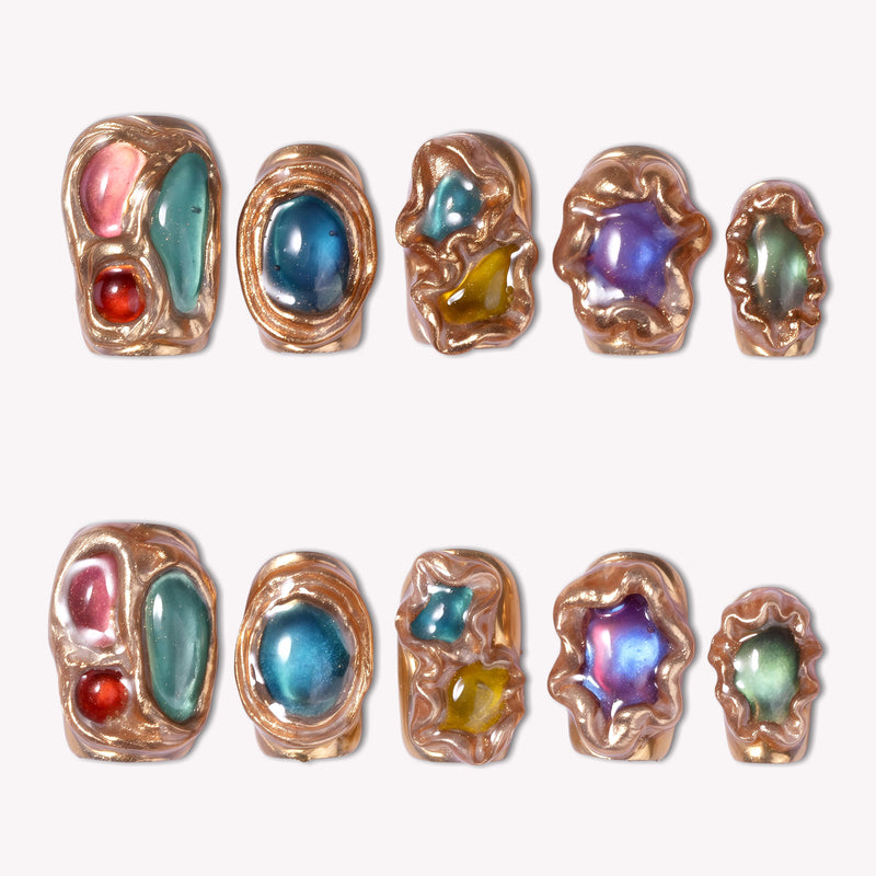 Luxury press-on nail art featuring sculptural metallic gold frames surrounding colorful gemstone charms in blue, purple, green, and amber tones, with chunky gold and black onyx rings, displayed against a neutral background