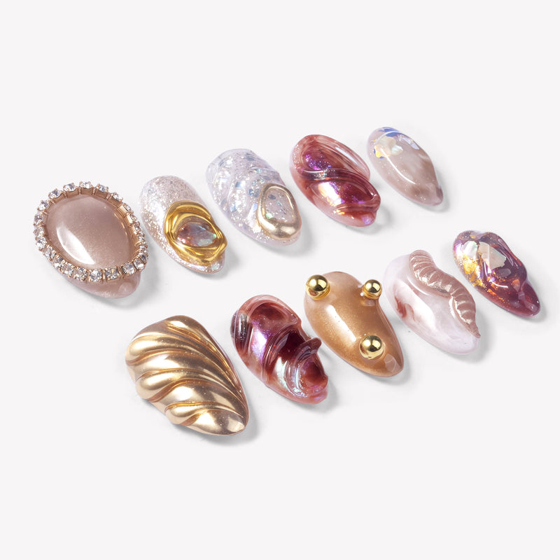 Angled flat lay of Molten Mirage press-on nails with amber and purple jelly swirls, champagne shimmer, gold shell accents, rhinestone trim, and raised gem details