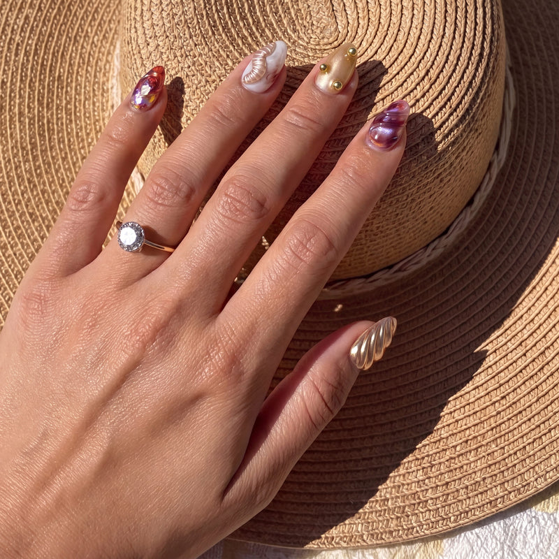 Hand model wearing Molten Mirage press-on nails on a straw hat with amber jelly swirls, gold shell accents, pearl details, and iridescent gem art