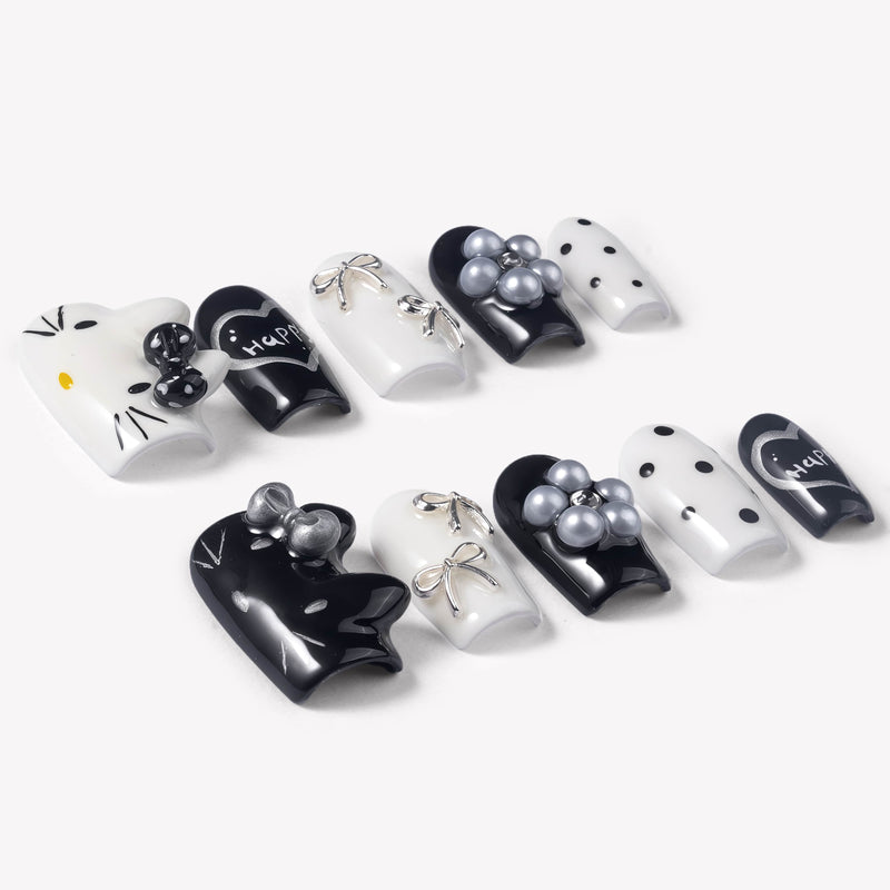 Angled flat lay of Monochrome Meow press-on nails with black and white cat designs, silver bow accents, pearl charms, polka dots, and happy heart lettering