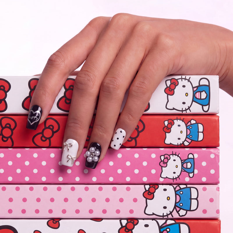 Hand model wearing Monochrome Meow press-on nails with black and white cat designs, silver bows, pearl charms, polka dots, and a colorful cartoon box background