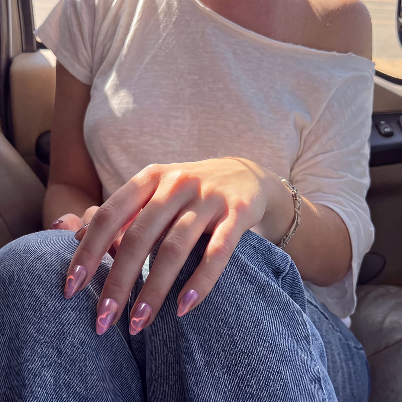 Nebula Kiss purple and pink cat-eye press-on nails with glowing hearts shown on a hand model wearing blue jeans
