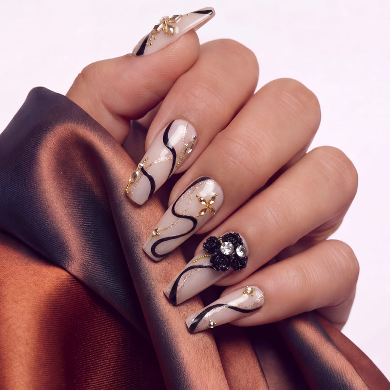 Close-up of a hand wearing beige press-on nails with black swirls, gold accents, and floral embellishments.