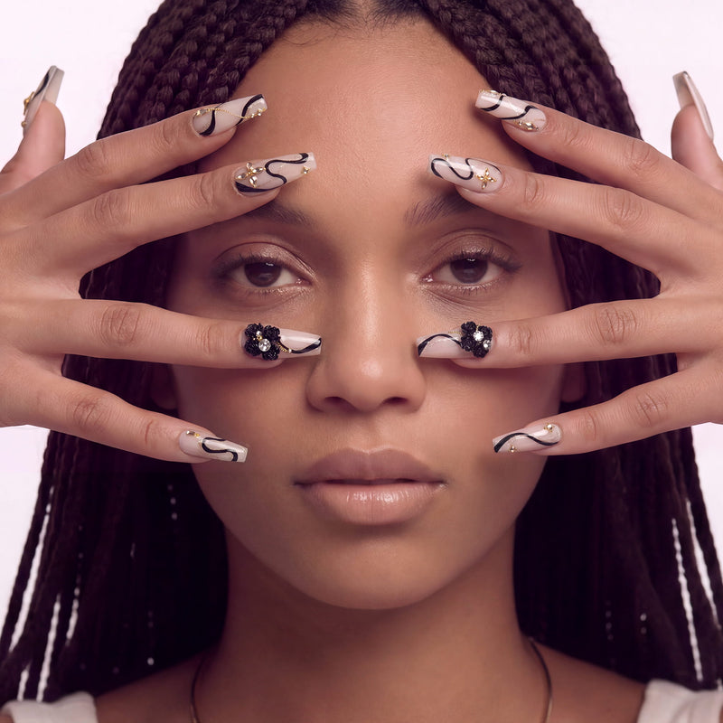 Hand model wearing beige press-on nails with black swirls, gold accents, and floral embellishments.