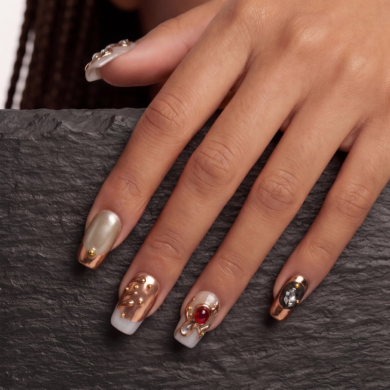 Hand model wearing pearl white and rose gold press-on nails with gold bead accents, a red jewel, and a black gemstone on a dark textured background.