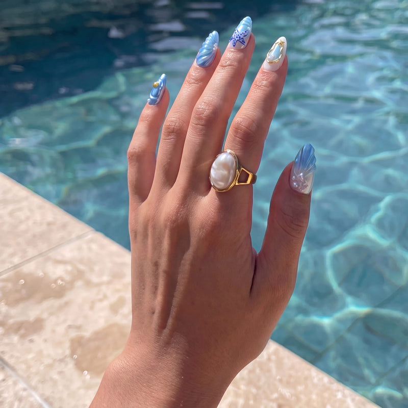 Hand displaying Ocean Rhapsody blue ocean-inspired press-on nails with shell and starfish details beside a swimming pool