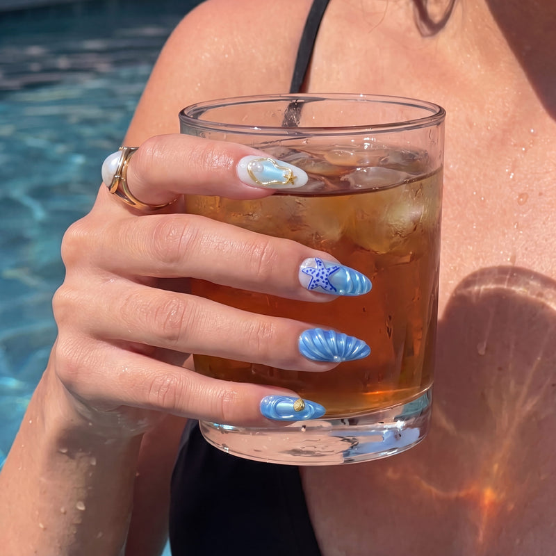 Hand wearing Ocean Rhapsody blue seashell and starfish press-on nails while holding a drink beside a pool