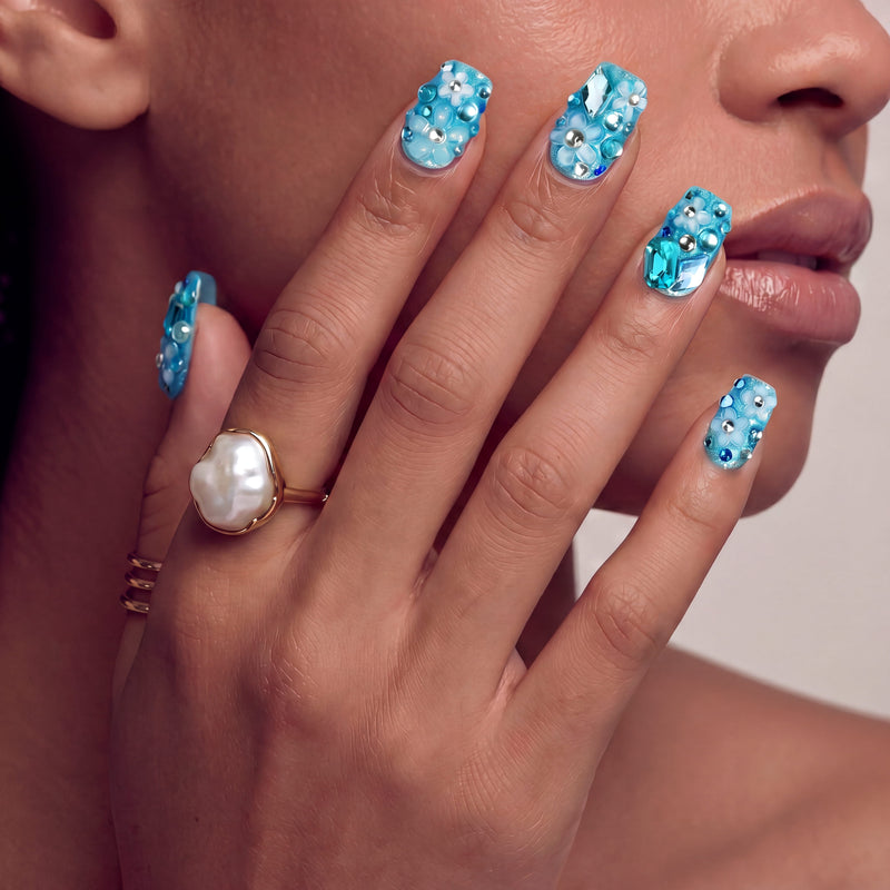 Hand model wearing Ocean Sparkle Bloom handmade press-on nails with blue rhinestones and a pearl ring.