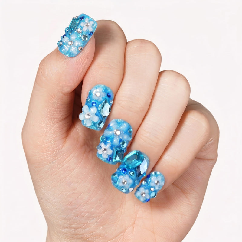 Close-up of a hand wearing blue rhinestone press-on nails on a white background.