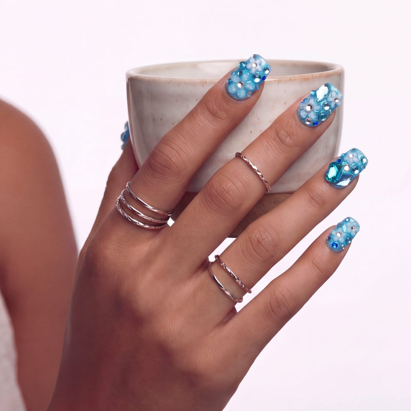 Hand model holding a ceramic cup while wearing blue rhinestone press-on nails and silver rings.