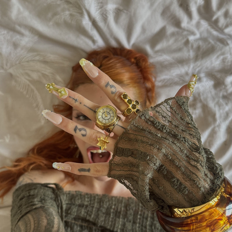 Face close-up hand model wearing Opal Dynasty press-on nails with nude pink bases, gold crown accents, pearl flowers, rhinestones, and rings