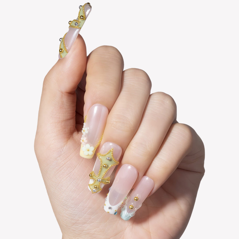 Hand model wearing Opal Dynasty press-on nails with nude pink bases, gold crown accents, pearl flower details, rhinestones, and blue translucent tips