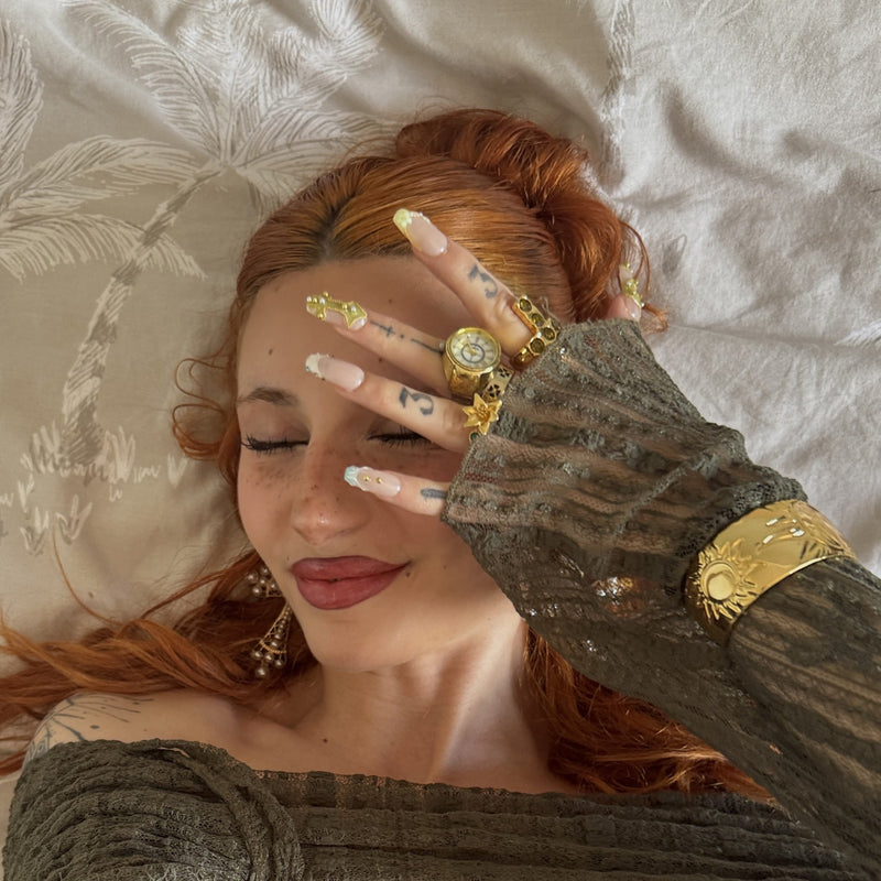 Reclining face close-up hand model wearing Opal Dynasty press-on nails with nude pink bases, gold accents, pearl details, rhinestones, and rings