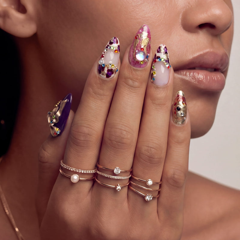 Close-up of a hand model wearing Opulent Ribbon Reverie press-on nails in purple pink and ivory with gold ribbon details and colorful gem accents