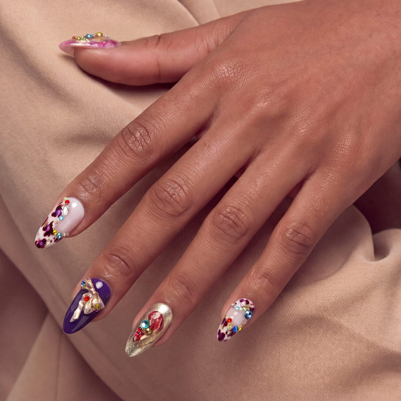 Hand model wearing Opulent Ribbon Reverie press-on nails in purple pink and ivory with gold ribbon details and colorful gem accents
