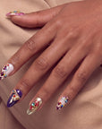 Hand model wearing Opulent Ribbon Reverie press-on nails in purple pink and ivory with gold ribbon details and colorful gem accents