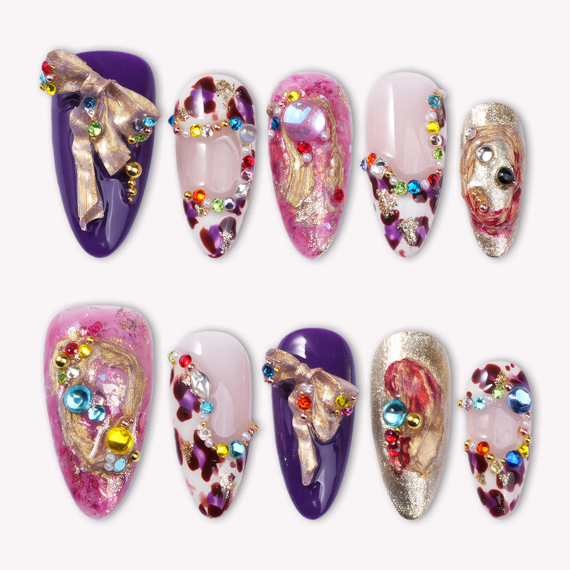 Opulent Ribbon Reverie press-on nails in purple pink and ivory with gold ribbon details and colorful gem accents arranged on a white background