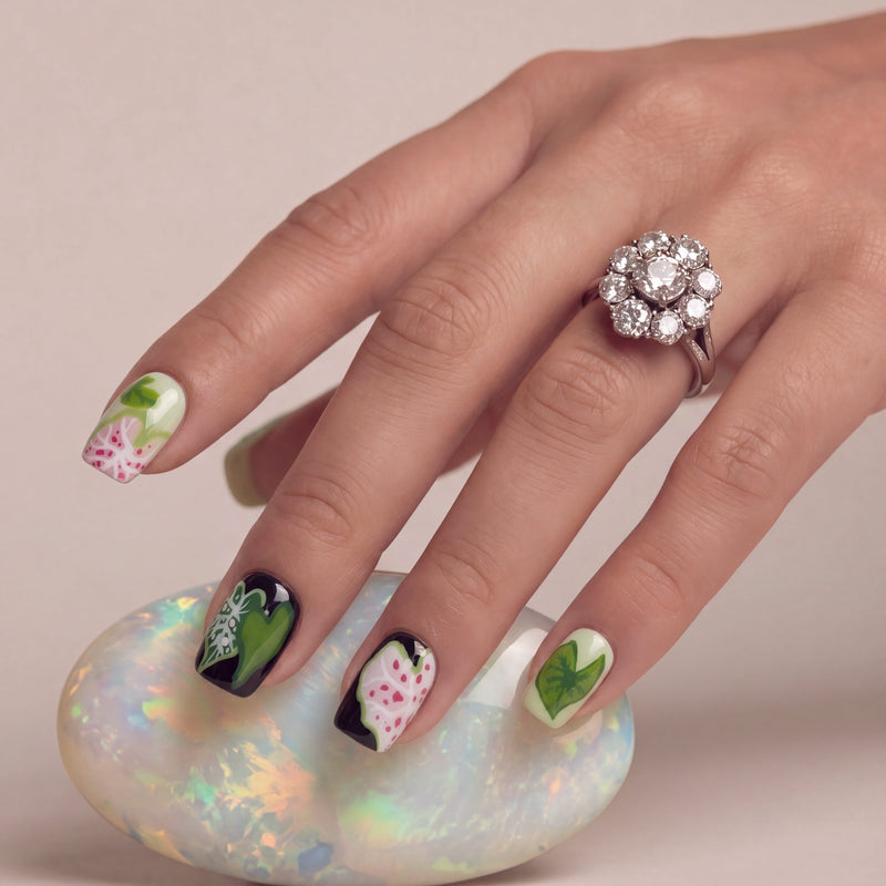 Hand model wearing Pink Caladium press-on nails with a floral leaf design, shown with a jeweled ring and iridescent stone prop