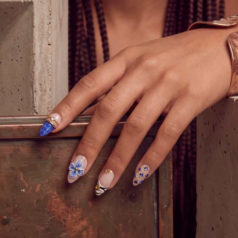 Hand model wearing Porcelain Azure handmade press-on nails with blue floral accents, gold beads, and a cobalt blue accent nail in a lifestyle pose.