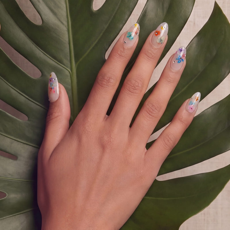 Hand model wearing floral press-on nails posed against a large green leaf.