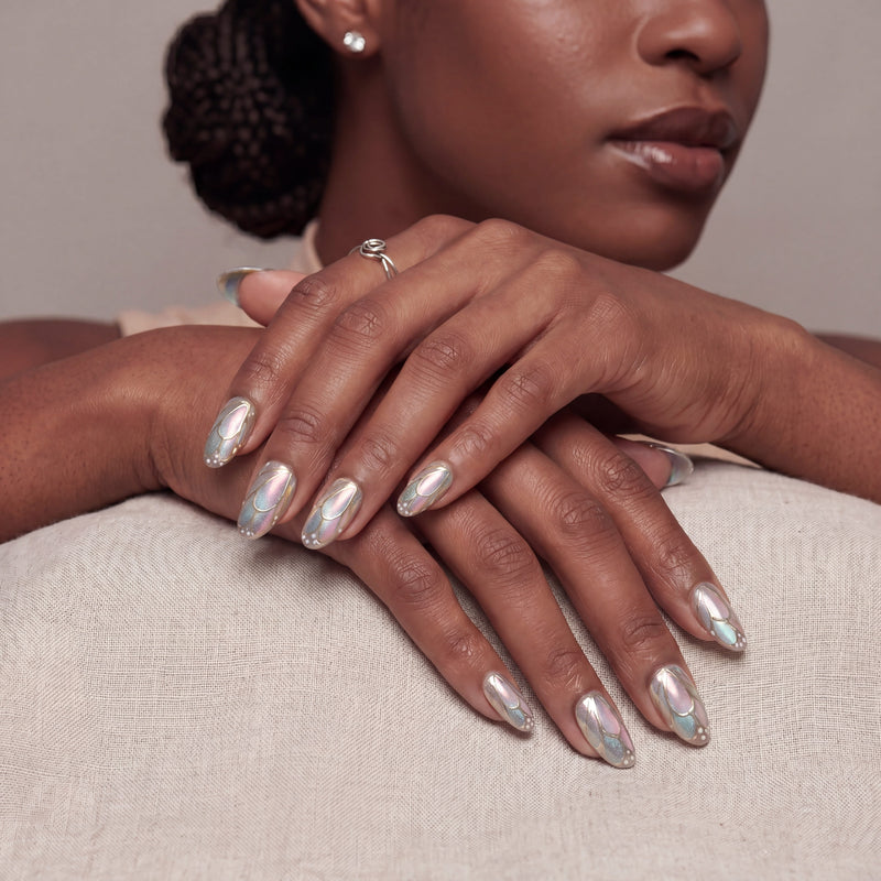 A hand model wearing iridescent press-on nails while resting both hands on a beige surface