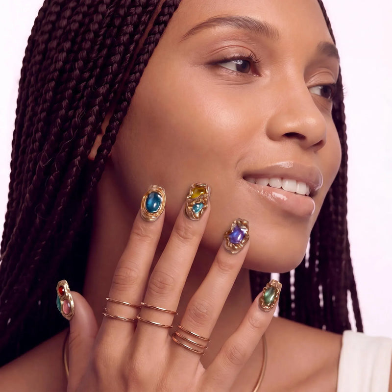 Close-up of a hand model wearing multicolor gemstone press-on nails with gold rings and a neutral background.