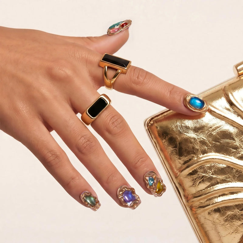 Hand model wearing multicolor gemstone press-on nails with gold rings beside a metallic gold clutch.