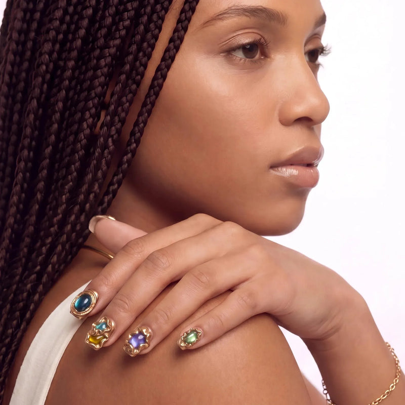 Shoulder close-up of a hand model wearing multicolor gemstone press-on nails with gold accents.