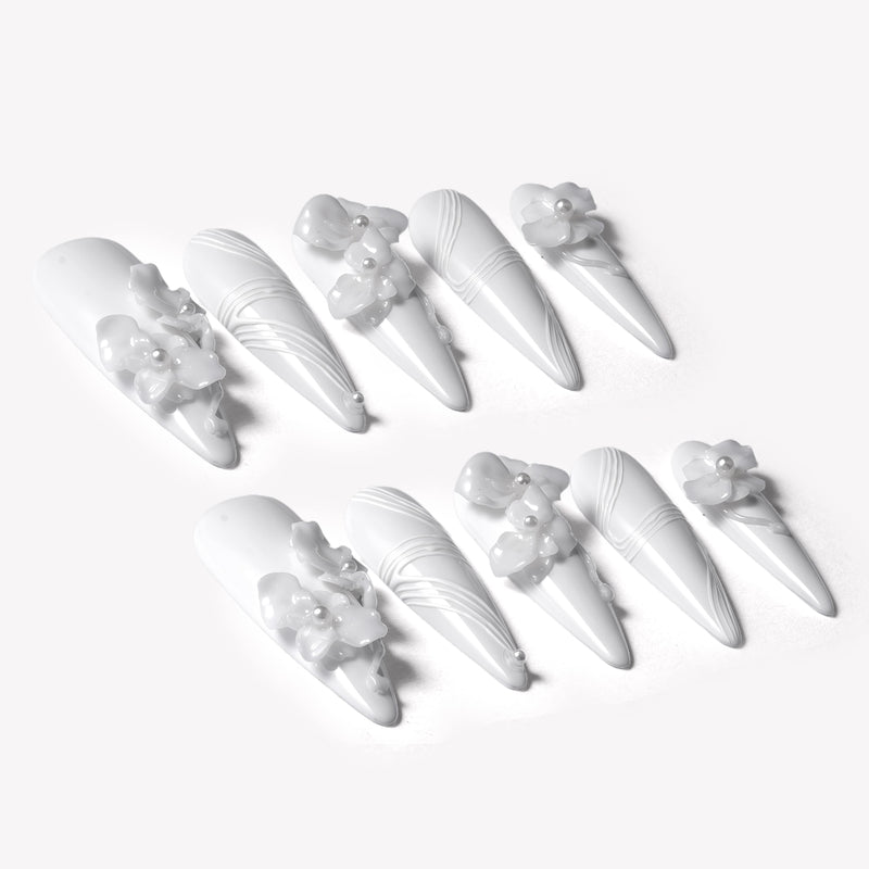 Angled flat lay of Quiet Sculpture press-on nails with glossy white stiletto shapes, raised 3D flowers, pearl accents, and sculpted line textures