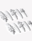Angled flat lay of Quiet Sculpture press-on nails with glossy white stiletto shapes, raised 3D flowers, pearl accents, and sculpted line textures