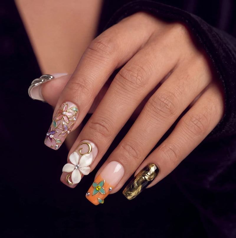 Hand model wearing ornate press-on nails with floral, gold, and sculpted details on a dark background.