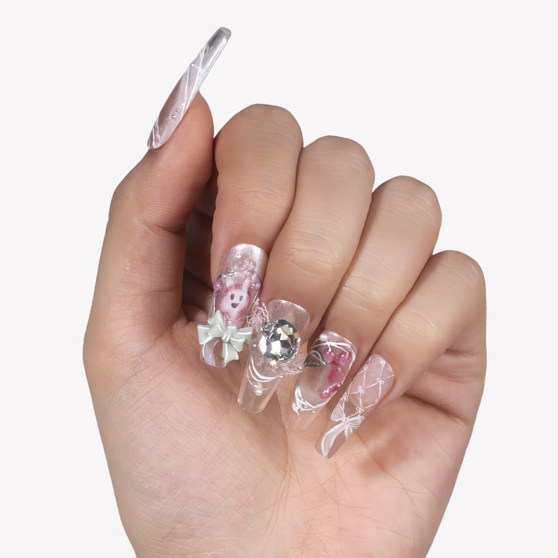 Hand model wearing Rococo Reverie press-on nails with clear pink bases, a rhinestone centerpiece, bow charm, pearls, and rose details
