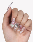 Hand model wearing Rococo Reverie press-on nails with clear pink bases, a rhinestone centerpiece, bow charm, pearls, and rose details