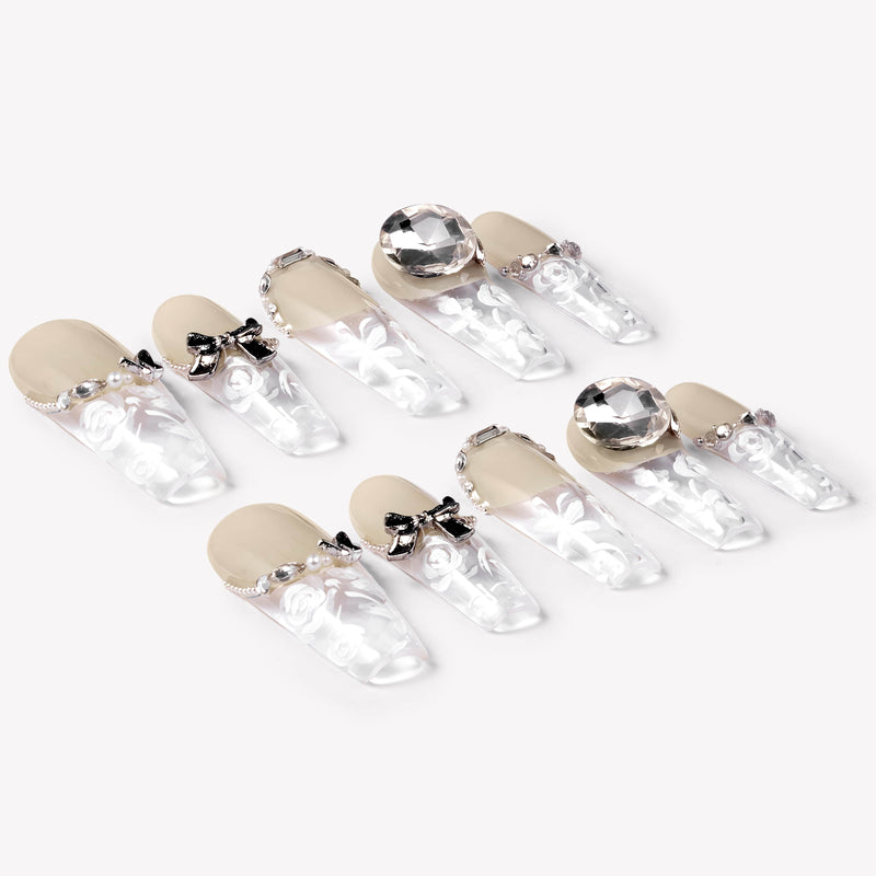 Angled flat lay of Rose Gold Whispers press-on nails with cream nude tops, translucent white floral tips, bow charms, rhinestone chains, and large crystals