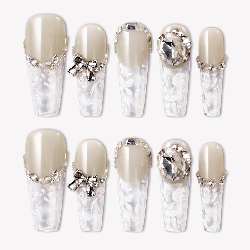 Rose Gold Whispers press-on nails with cream nude tops, translucent white floral tips, rhinestone chains, a bow charm, and large crystal accents