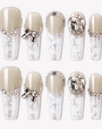Rose Gold Whispers press-on nails with cream nude tops, translucent white floral tips, rhinestone chains, a bow charm, and large crystal accents
