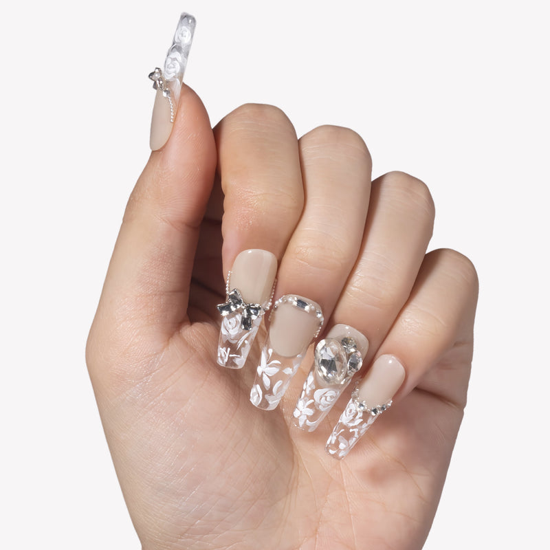 Hand model wearing Rose Gold Whispers press-on nails with cream nude bases, translucent white floral tips, a bow charm, and crystal rhinestones