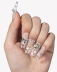 Hand model wearing Rose Gold Whispers press-on nails with cream nude bases, translucent white floral tips, a bow charm, and crystal rhinestones