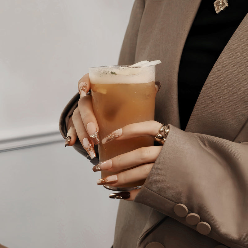Lifestyle hand model in a beige blazer holding a drink while wearing Rosegold Flutter press-on nails with rosegold butterfly and rhinestone details