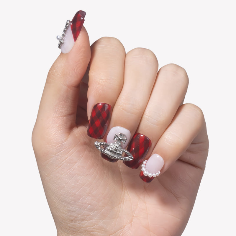 Hand model wearing Royal Rebellion press-on nails with red black argyle pattern, white gradient accents, silver orb charm, and pearl details
