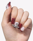 Hand model wearing Royal Rebellion press-on nails with red black argyle pattern, white gradient accents, silver orb charm, and pearl details