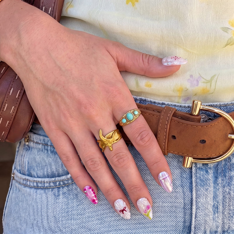 Hand model wearing Sakura Whisper pink and pearly white floral press-on nails with denim jeans, gold rings and a brown belt