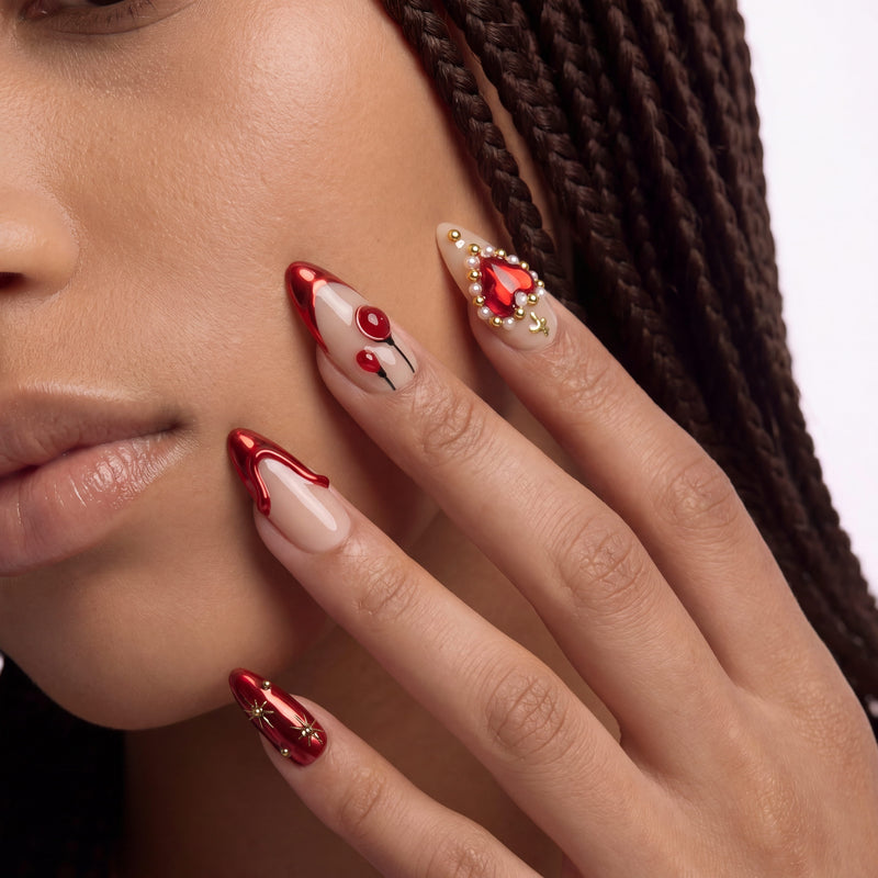 Close-up of a hand model wearing Scarlet Heart Oath press-on nails with red metallic tips, nude bases, hearts, and gold bead accents