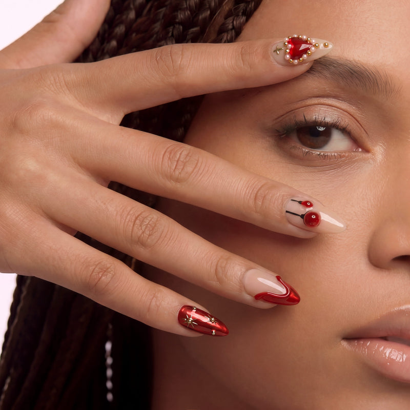 Lifestyle portrait of a hand model wearing Scarlet Heart Oath press-on nails with red metallic tips, nude bases, and gold bead heart accents