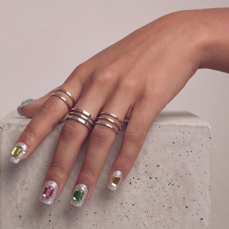 Hand model wearing silver holographic glitter press-on nails with large emerald-cut rhinestones in jewel tones.