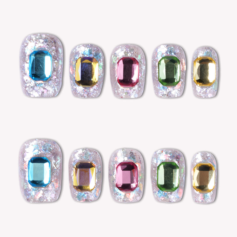 A set of 10 luxury press-on nails with a sparkling silver holographic glitter base, each featuring a large emerald-cut rhinestone in vibrant jewel tones including cyan blue, amber gold, magenta pink, emerald green, and champagne gold, displayed in two horizontal rows on a clean white background