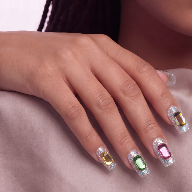 Hand model wearing silver holographic glitter press-on nails with emerald-cut rhinestones on a soft blush background.