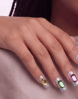 Hand model wearing silver holographic glitter press-on nails with emerald-cut rhinestones on a soft blush background.