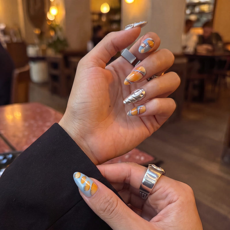 Cafe lifestyle view of Solar Pulse orange, pale blue and sculpted silver chrome press-on nails on a hand model